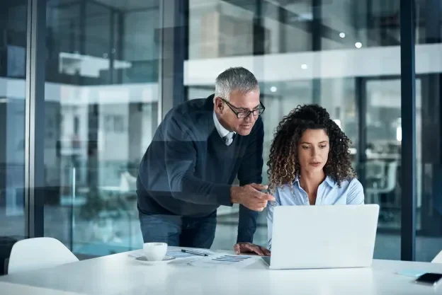 A man in glasses leans over a woman's shoulder to review a laptop with her