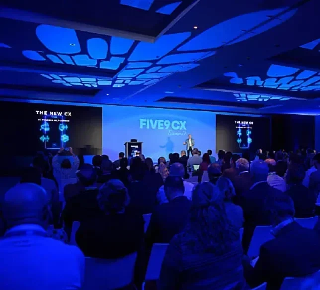 A speaker presents on stage to an audience in a dimly lit conference room.