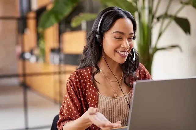 Smiling woman wearing a headset, engaging with a laptop in a bright, modern workspace.